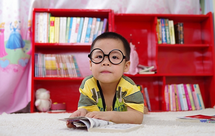 Kleiner Junge sitzt vor Bücherregal und blickt in die Kamera. Vor ihm liegt ein Buch. Auf seiner Nase trägt er eine auffällige Brille