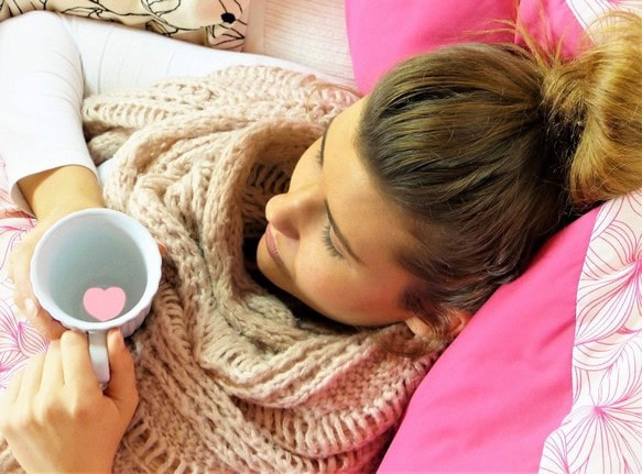 Eine kranke Person ruht sich mit einer leeren Tasse in der Hand im Bett aus.