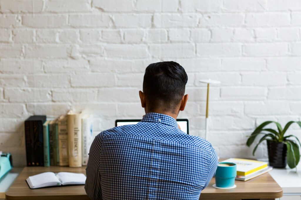 Rückenansicht eines Mannes, der an einem Schreibtisch sitzt und auf einen Laptop schaut.